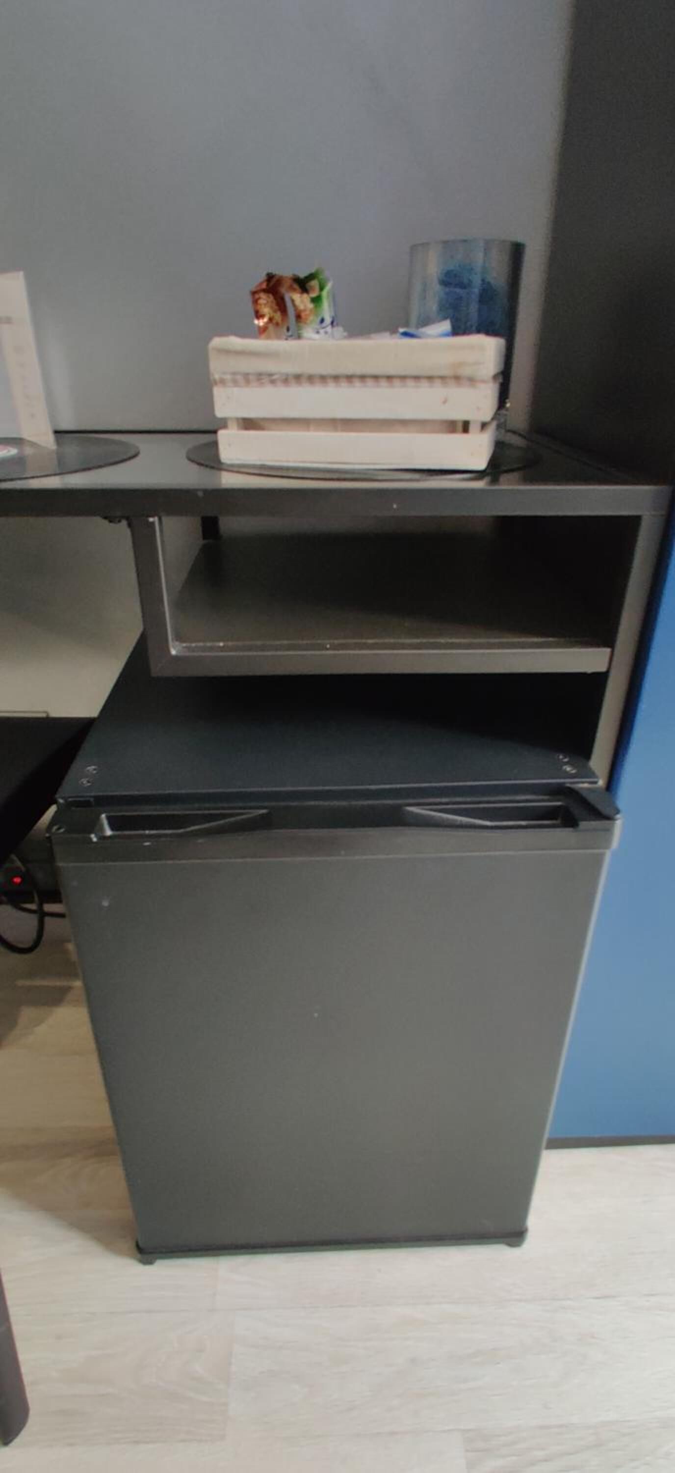 Un refrigerador compacto de color oscuro con un compartimento congelador en la parte superior, ubicado en una habitación con piso de color claro y pared azul.
