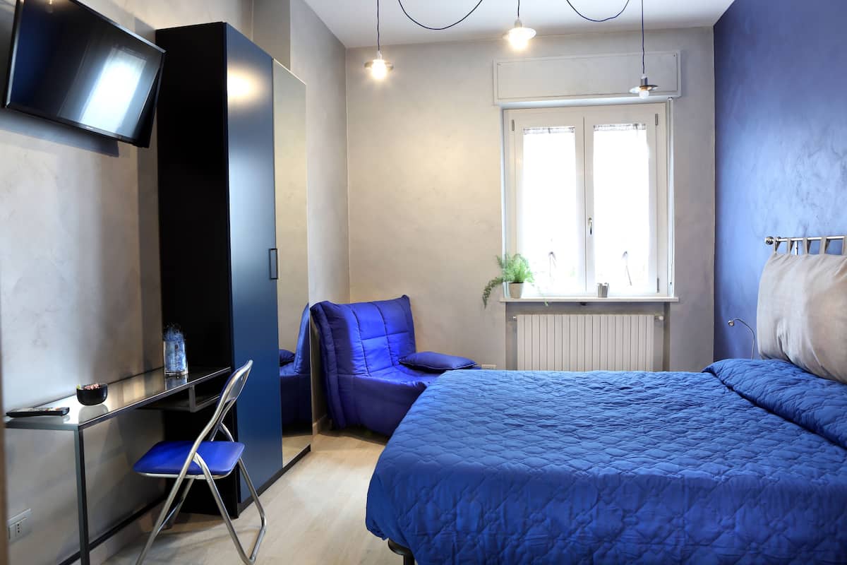 A modern bedroom with a large bed, a desk with a chair, a TV mounted on the wall, and a window with a view of the outdoors.