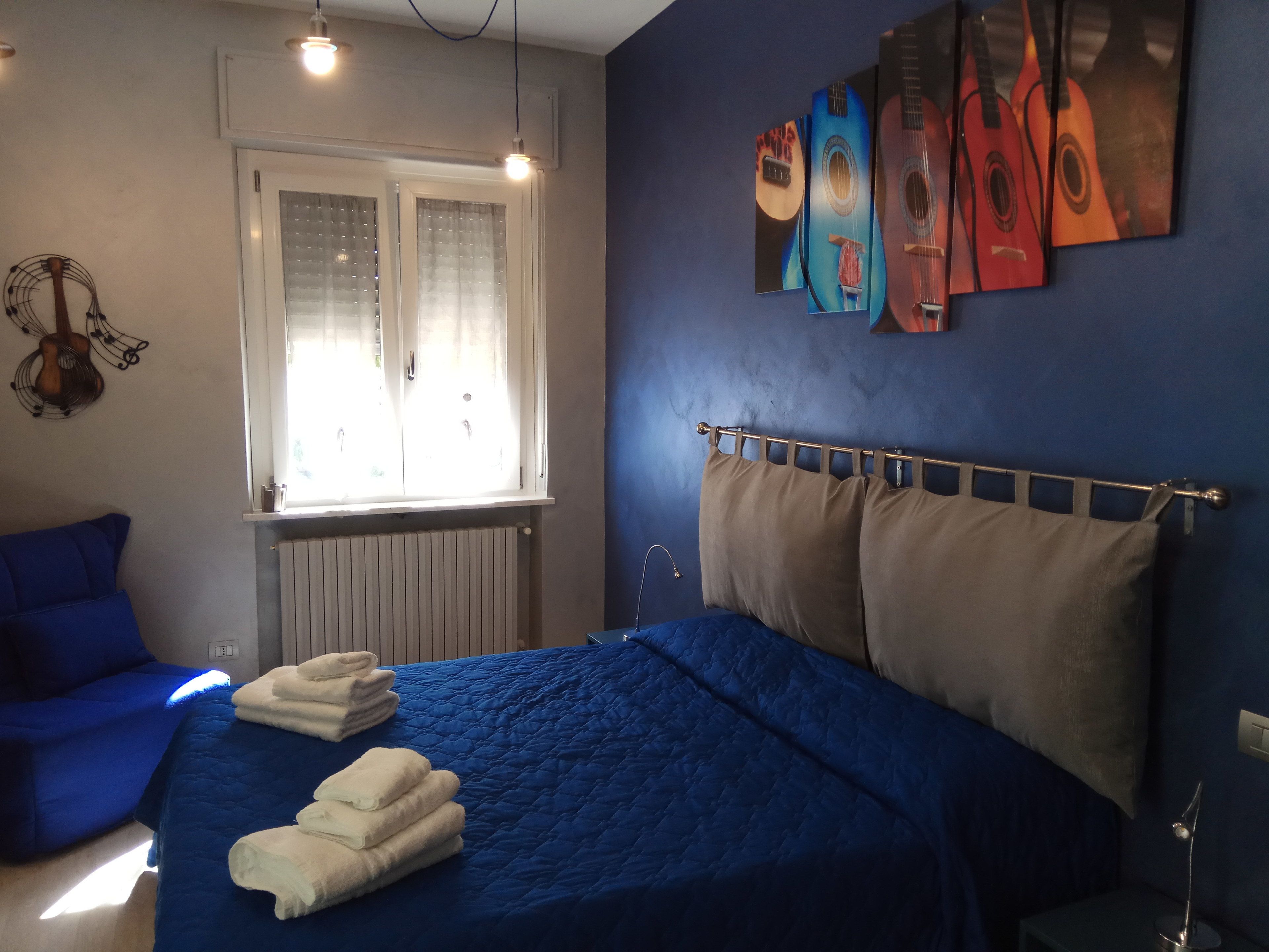 A bedroom with a blue bedspread, a window with blinds, a guitar wall decoration, and a headboard with multiple pillows.