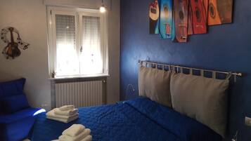 A bedroom with a blue bedspread, a window with blinds, a guitar wall decoration, and a headboard with multiple pillows.