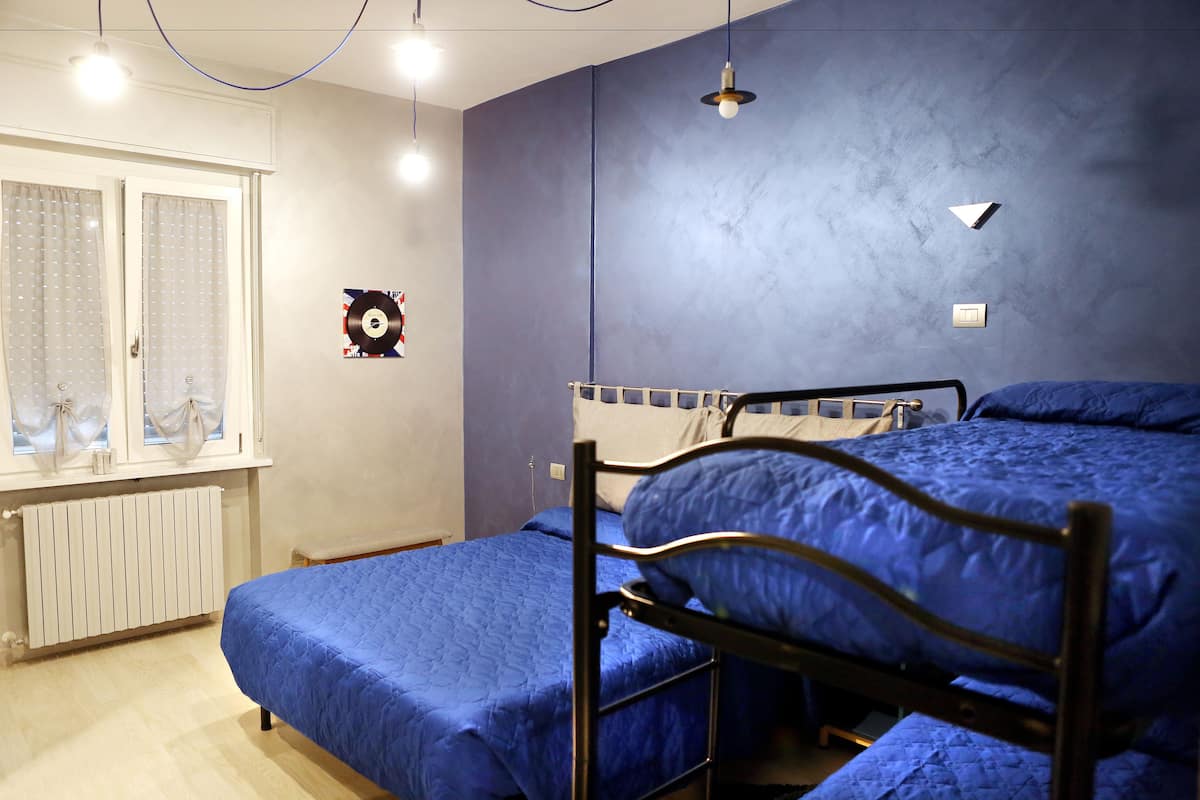 A room with a bunk bed, a single bed, a radiator, a window with curtains, and a wall-mounted light fixture.