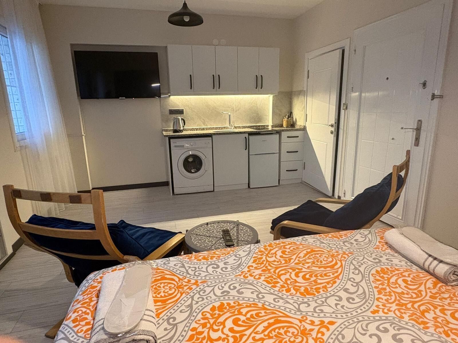 Habitación de hotel compacta con una cama, dos sillas, una mesita, lavadora y kitchenette.
