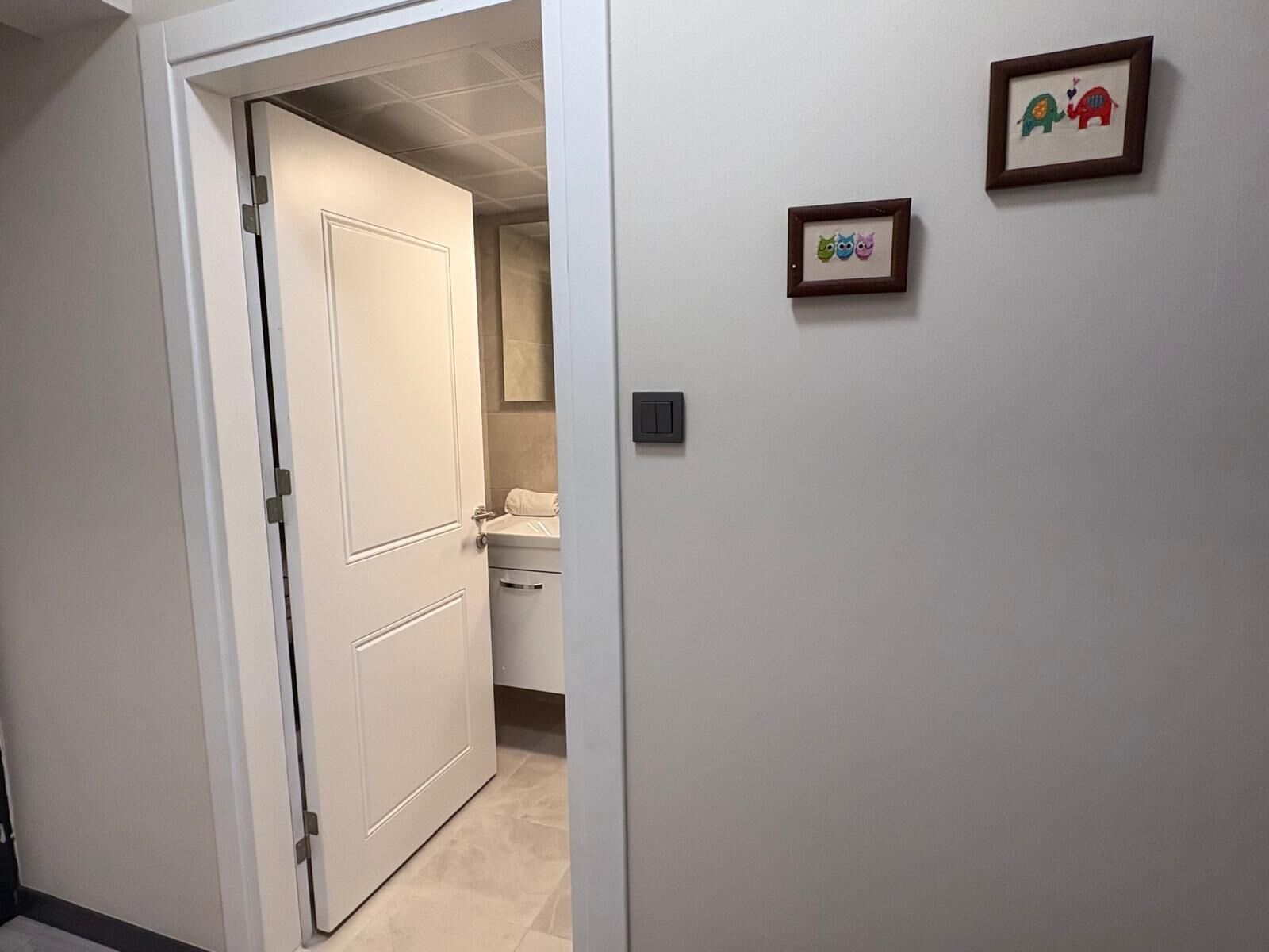 A hallway with a white door leading to a bathroom with a toilet and sink. There are two framed pictures on the wall.