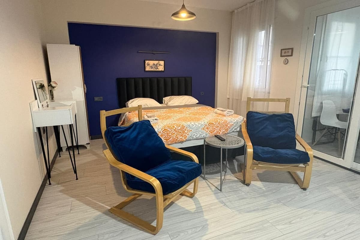 Quarto com cama, duas cadeiras azuis, uma mesinha, mesa de cabeceira, espelho, quadro na parede e porta de vidro deslizante.