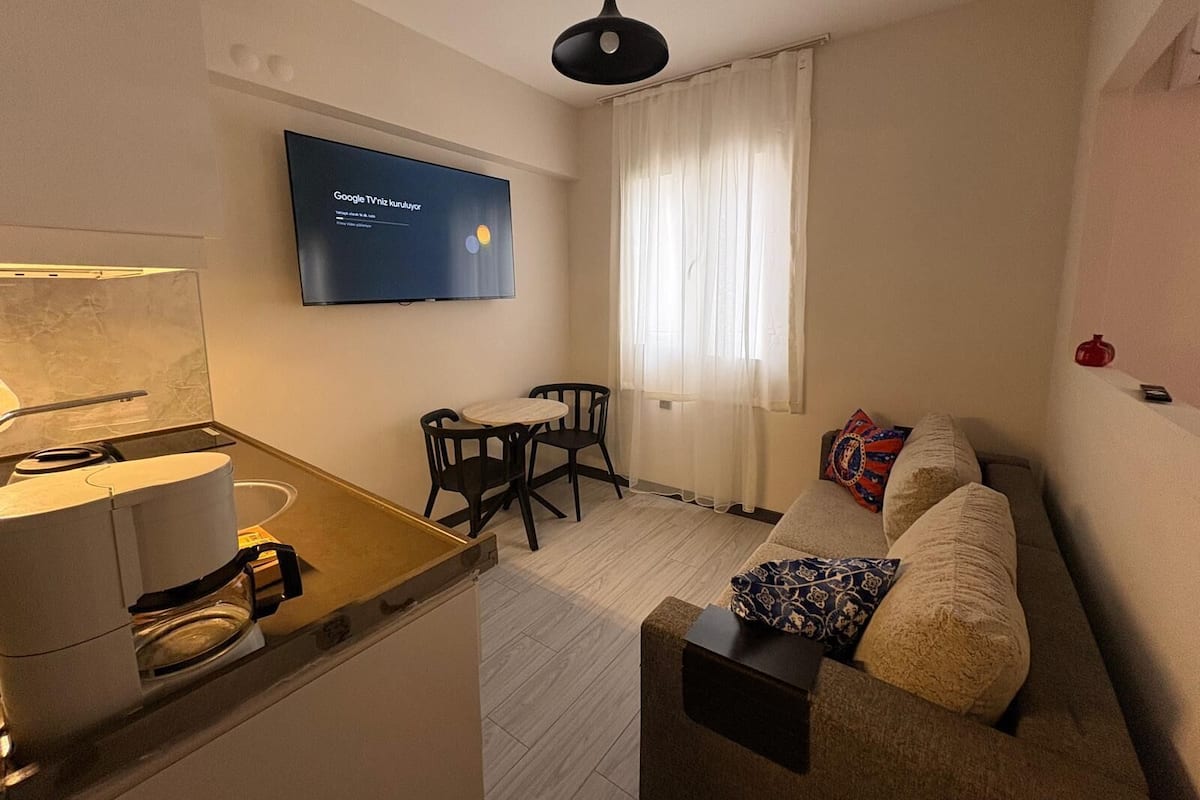 Um espaço compacto com cozinha compacta, TV de tela plana na parede, sofá com almofadas estampadas e área de refeições com mesa e cadeiras.