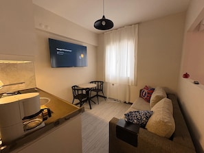 A compact living space with a kitchenette, a flat-screen TV mounted on the wall, a sofa with patterned cushions, and a dining area with a table and chairs. - Deka Evleri (Izmir)