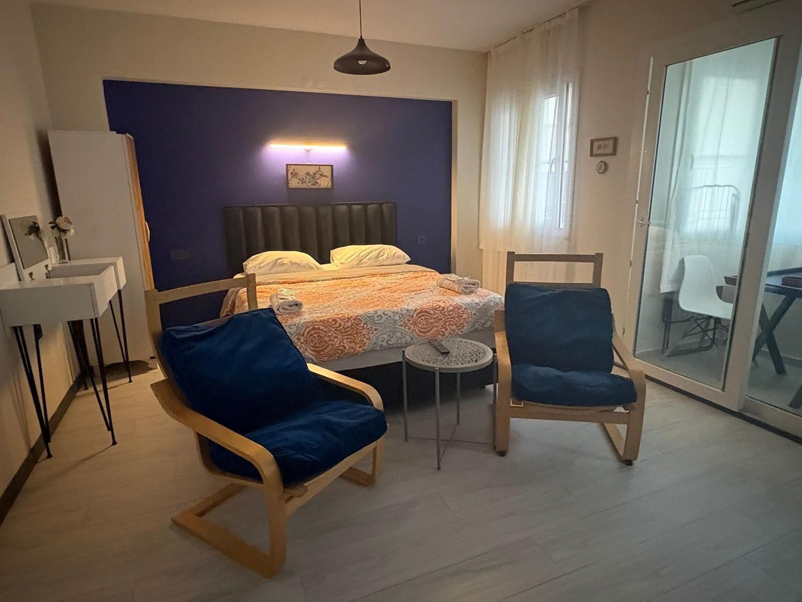 Een kamer met een bed, twee stoelen, een klein rond tafeltje, een bureau en een glazen deur die uitkomt op een balkon.