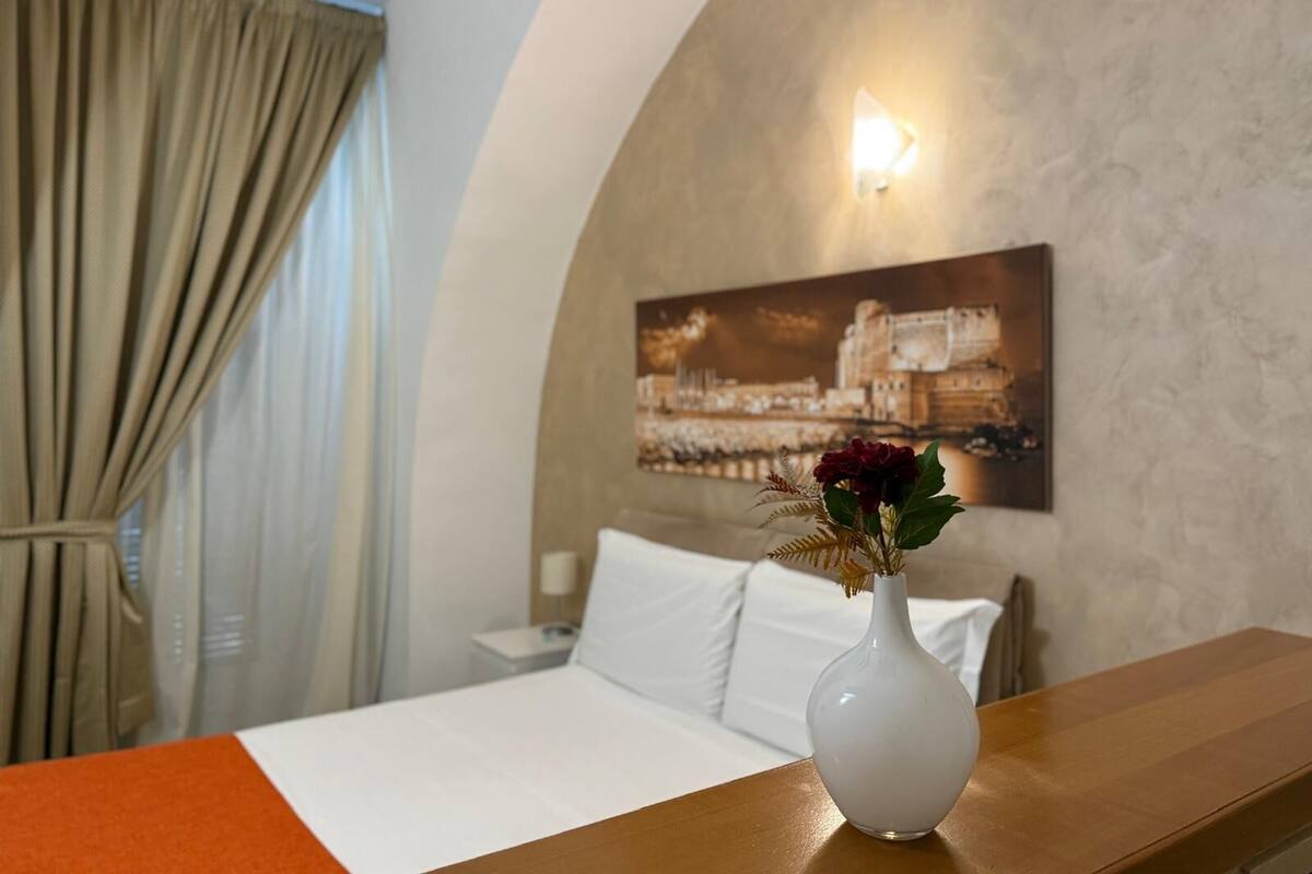 A hotel room with a bed, a white vase with flowers, a painting on the wall, and a window with curtains.