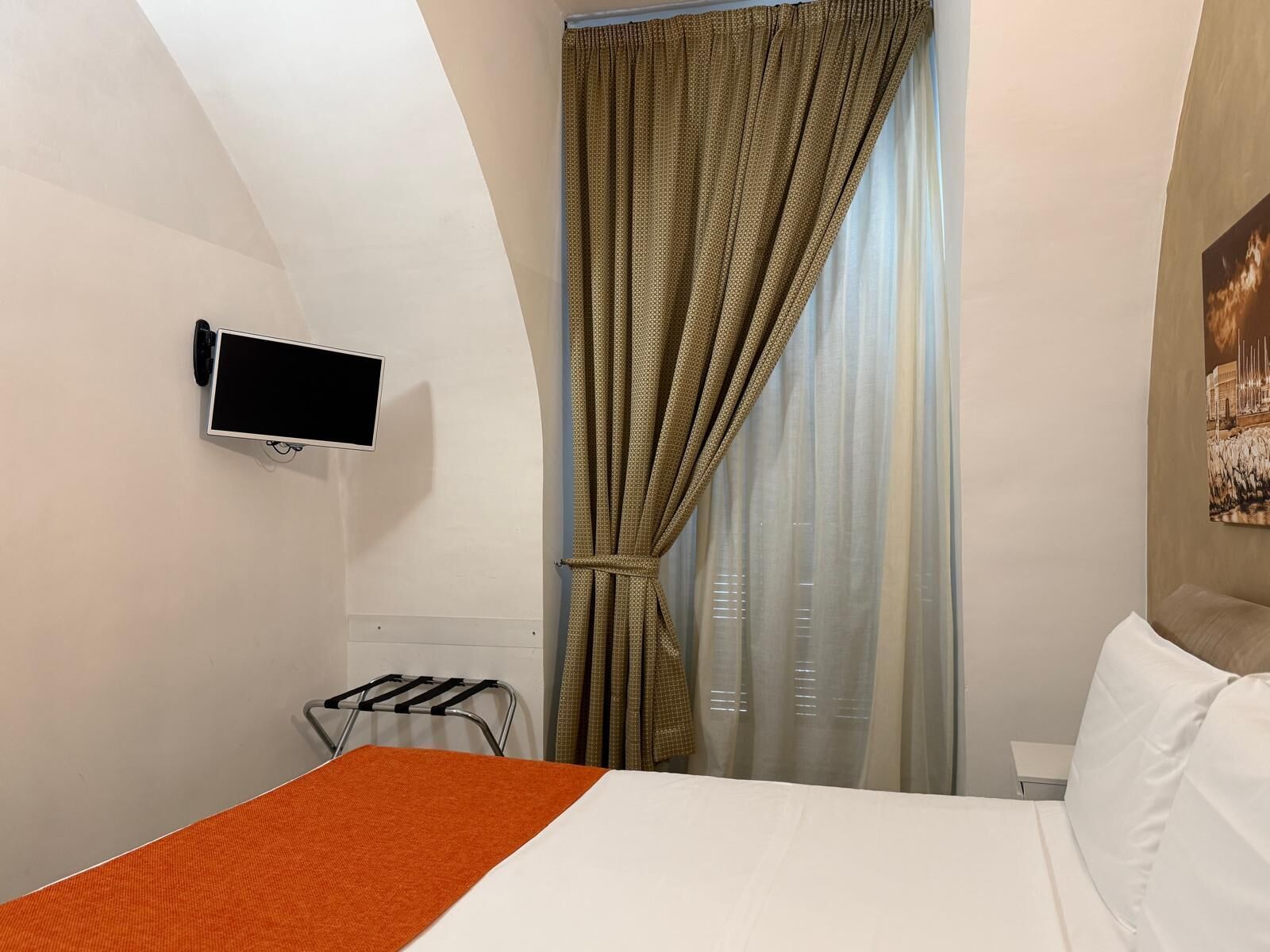 A hotel room with a bed, a TV mounted on the wall, a window with curtains, and a radiator.