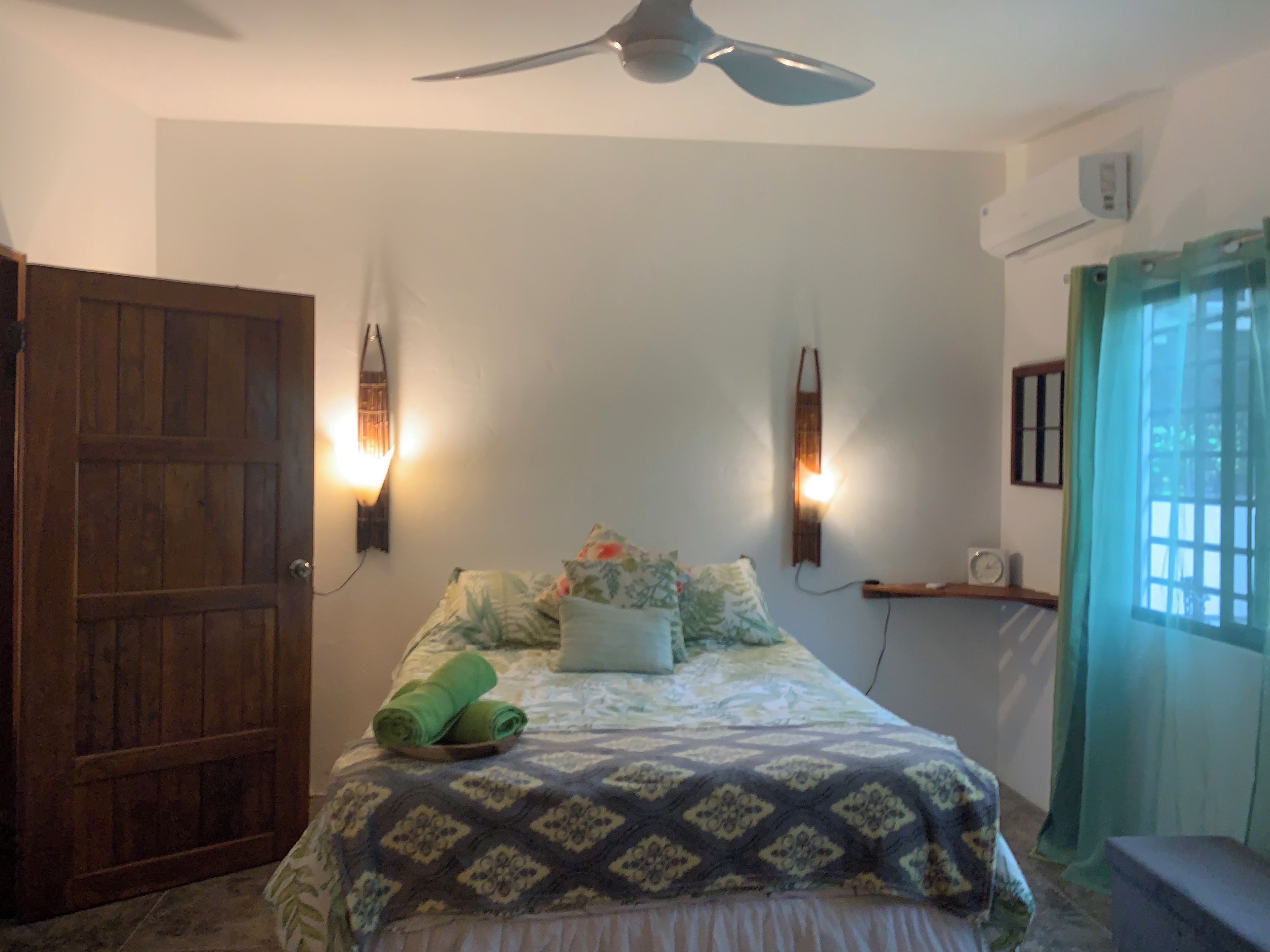 A bedroom with a bed, a ceiling fan, wall-mounted lights, a wooden door, and a window with curtains.