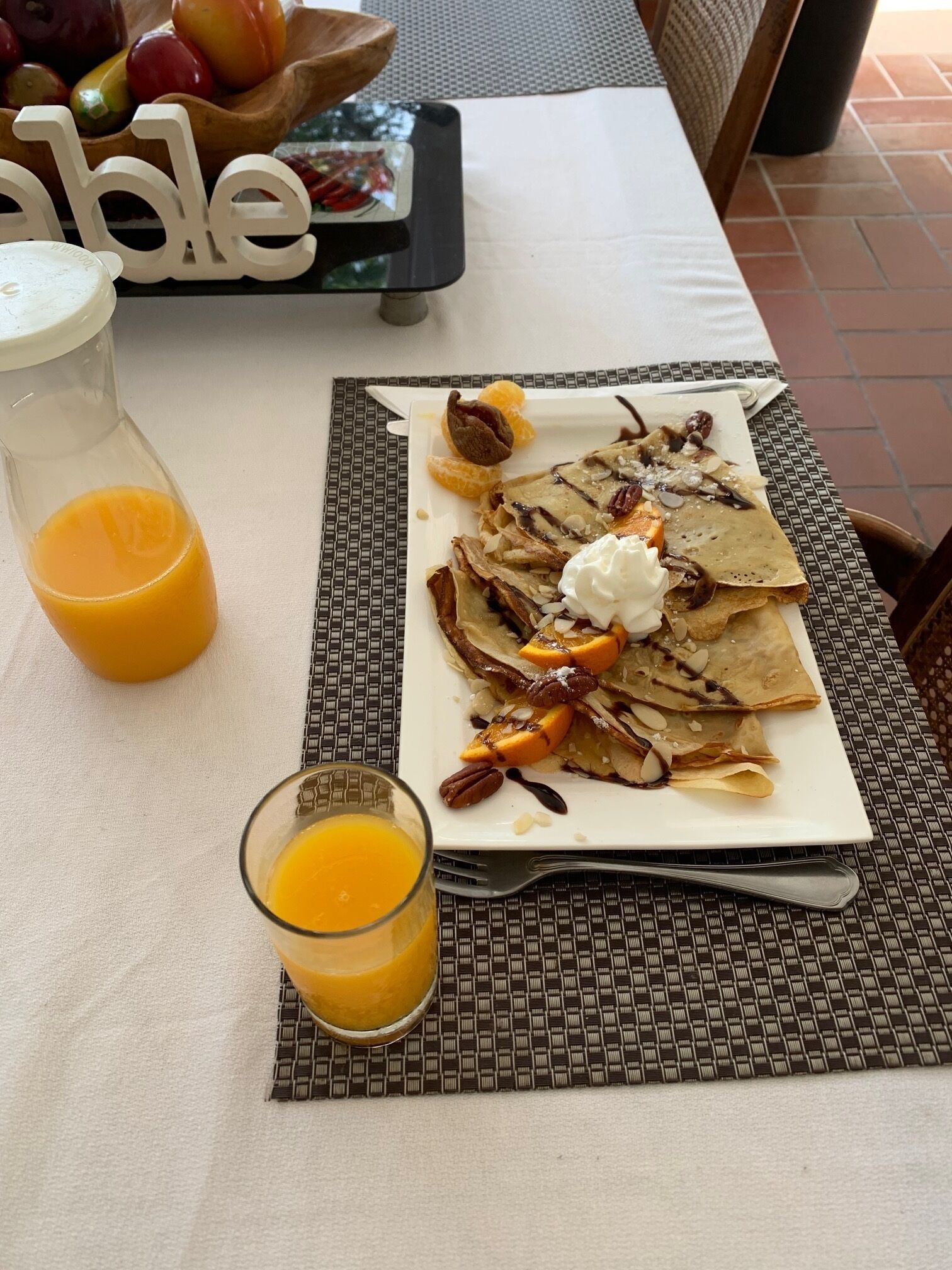 Un desayuno con una fuente de tortitas cubiertas de fruta, nueces y crema batida, acompañado de un vaso de zumo de naranja.