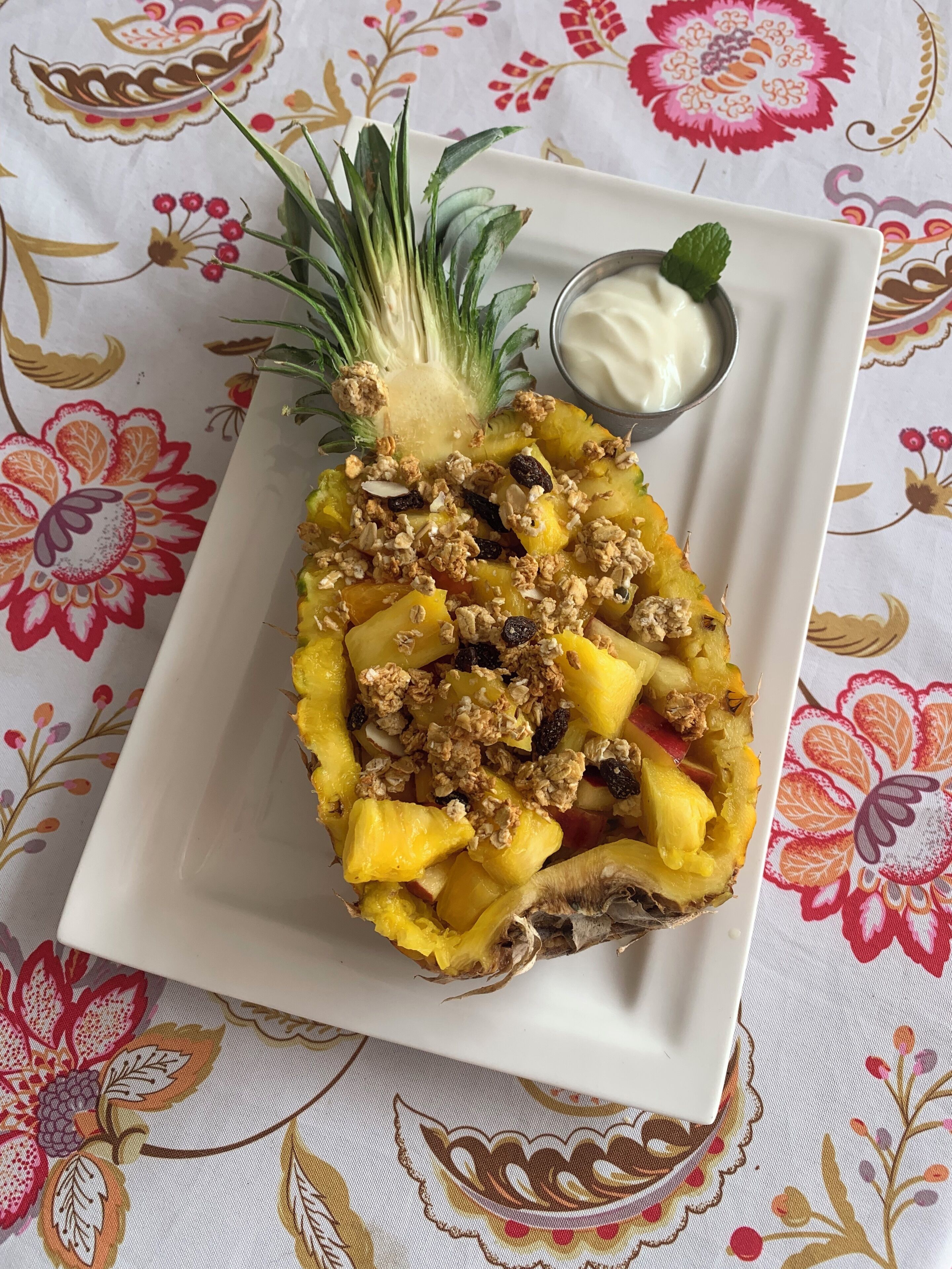 Una piña cubierta de granola y un tazón pequeño de yogur sobre un mantel estampado de flores.