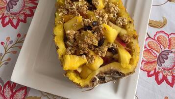 Una piña cubierta de granola y un tazón pequeño de yogur sobre un mantel estampado de flores.