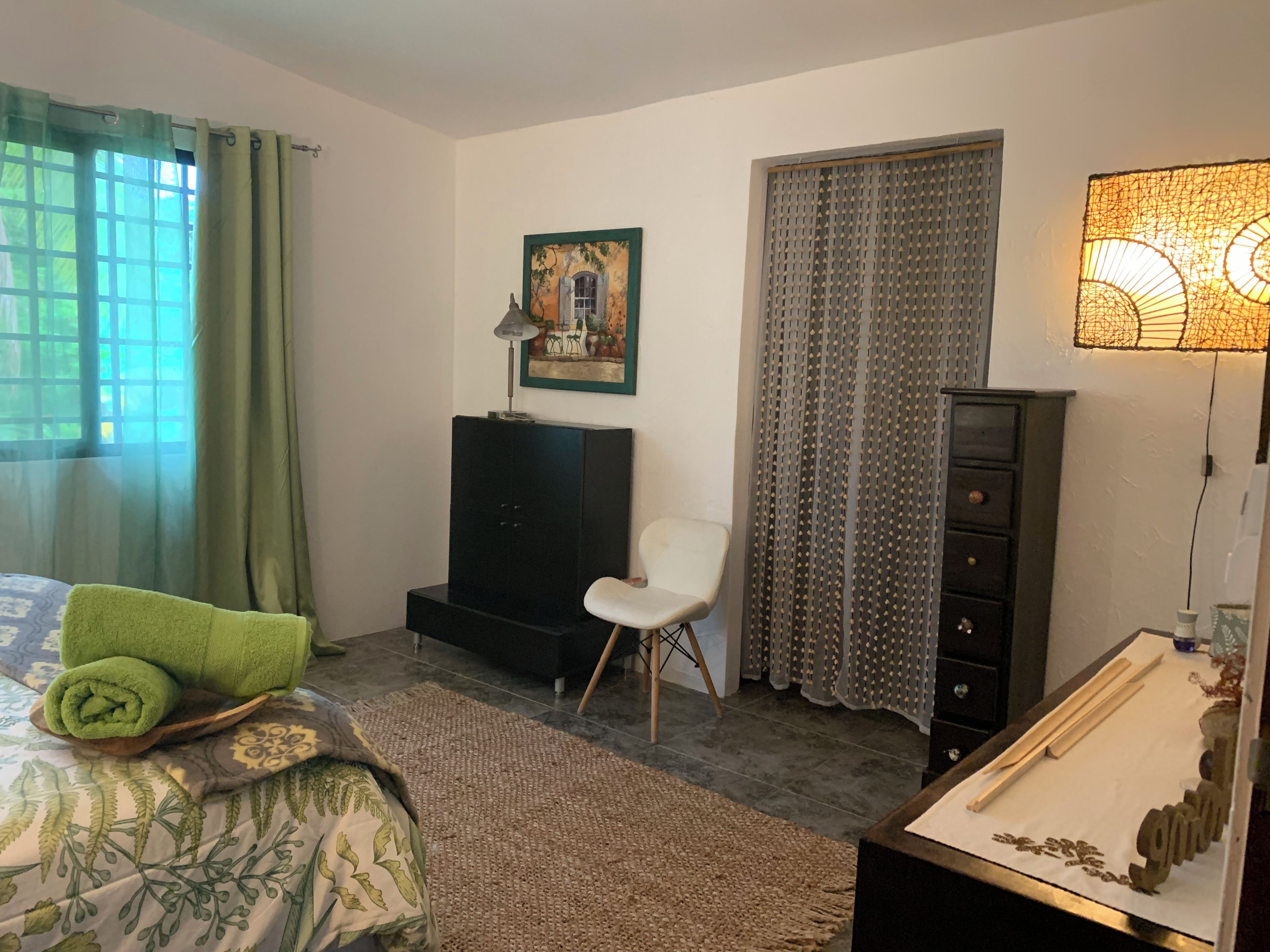 Una habitación con cama, una silla, escritorio, lámpara, cuadro y ventana con cortinas.