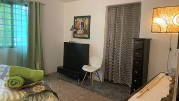 Una habitación con cama, una silla, escritorio, lámpara, cuadro y ventana con cortinas.