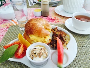 Un plato de desayuno con un croissant, huevos revueltos, tortitas de patata, una rebanada de sandía, un pequeño bol de yogur con frutos secos y rodajas de tomate fresco.