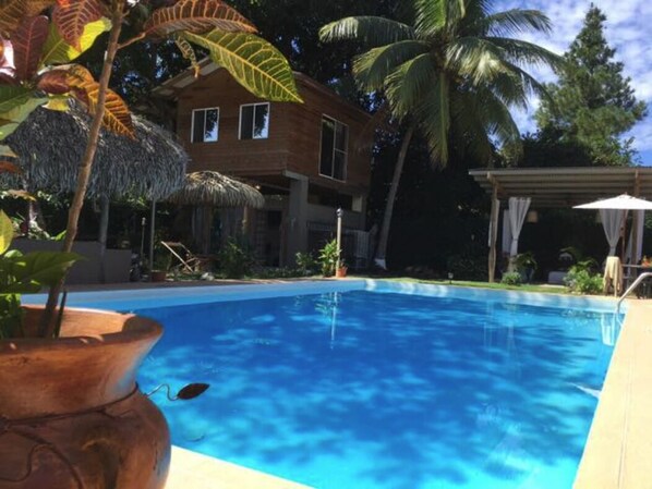 Una piscina azul cristalina rodeada por una casa de madera con techo de paja, palmeras y un espacio exterior cubierto.