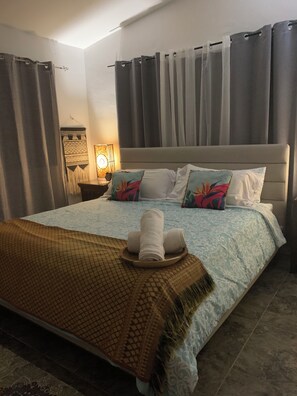 Una cama bien tendida con una colcha estampada, almohadas con diseños florales y una toalla enrollada sobre una bandeja.