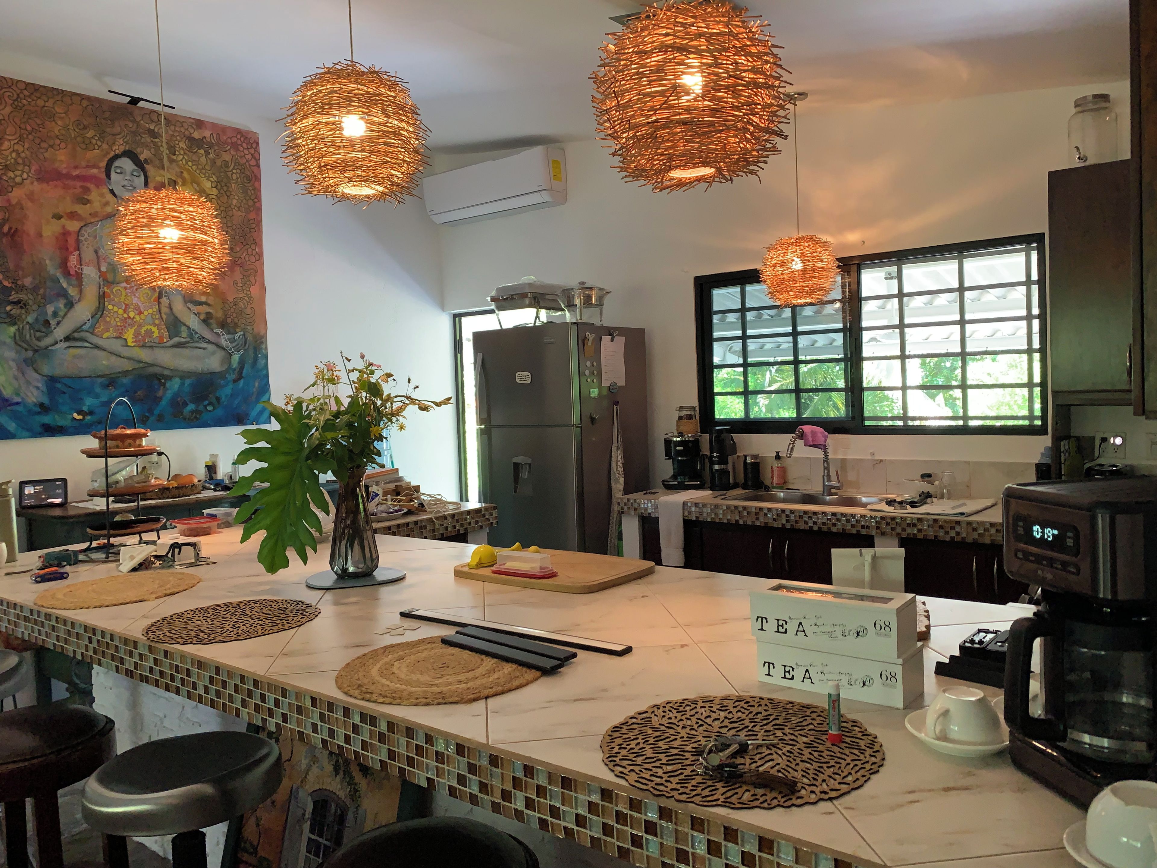 Una cocina con una isla grande, lámparas colgantes de mimbre, un refrigerador, una cafetera y una ventana con vistas a la vegetación.