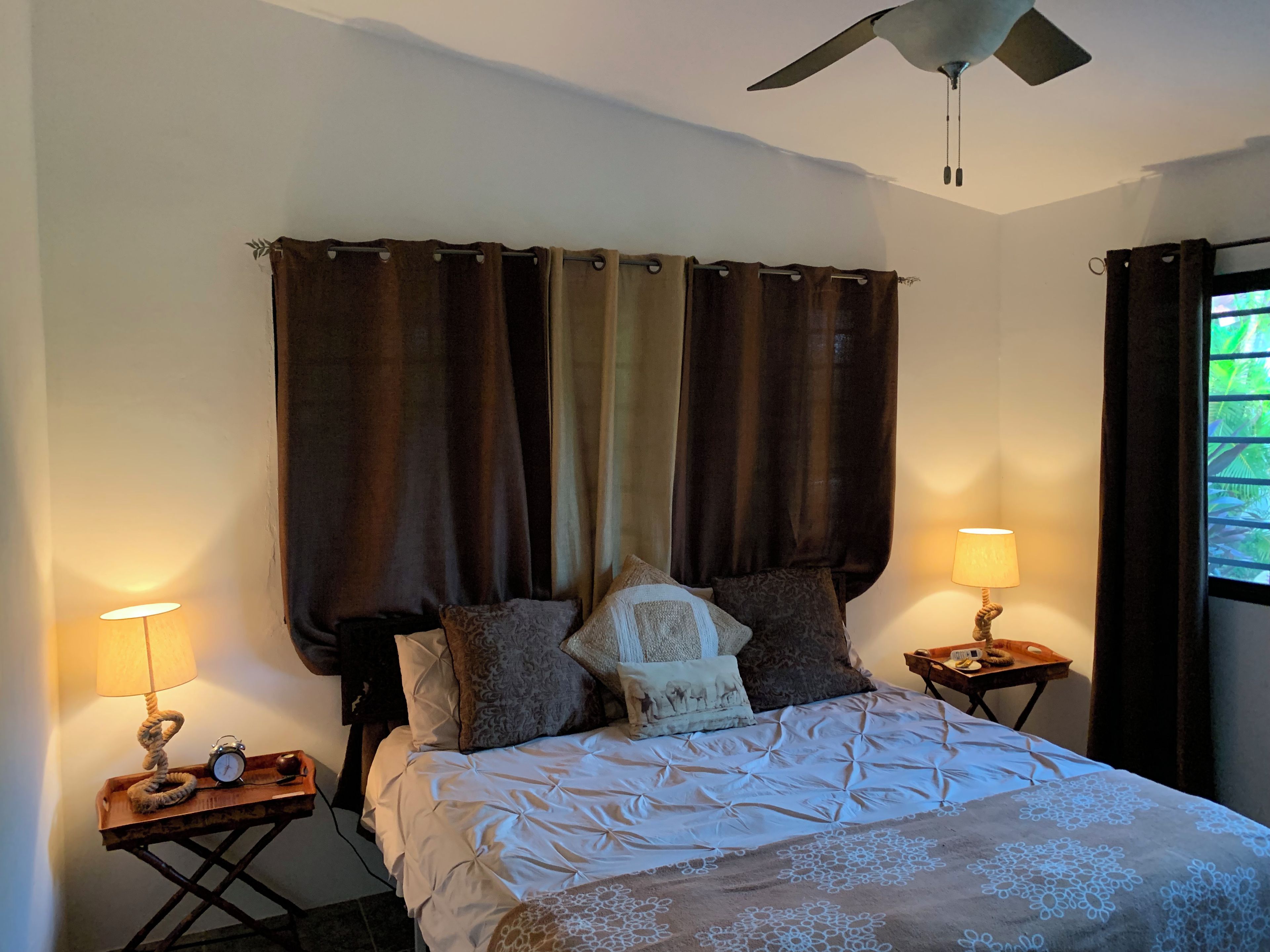 A bedroom with a bed, two bedside tables, a ceiling fan, and a window with curtains.