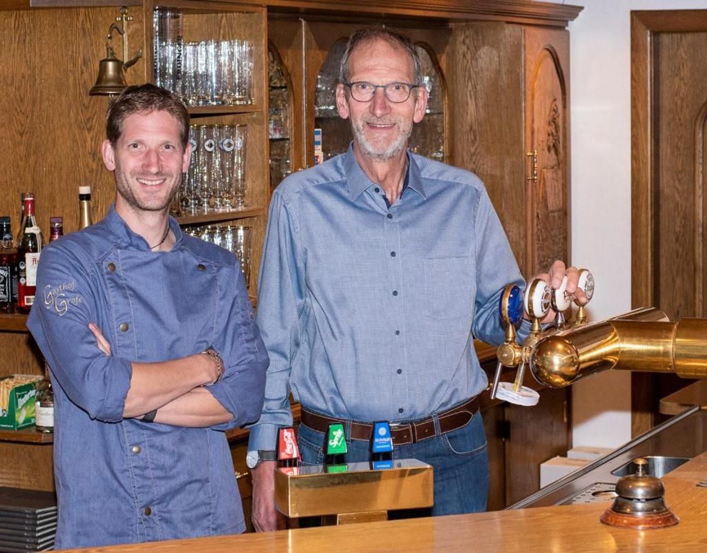 Two individuals standing in a bar area with a wooden cabinet in the background, a bar counter, and a brass beer tap.