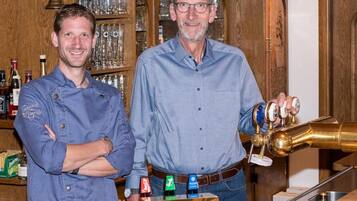 Two individuals standing in a bar area with a wooden cabinet in the background, a bar counter, and a brass beer tap.