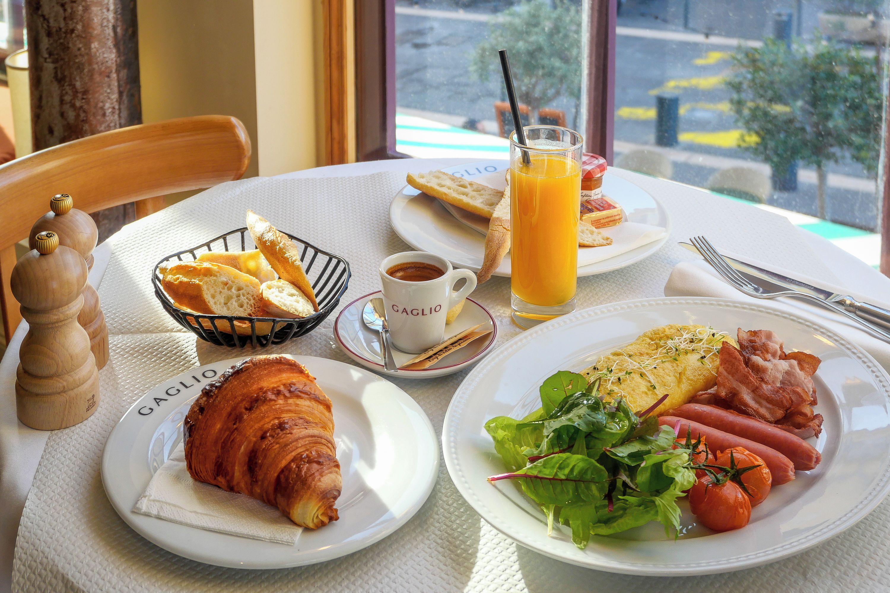 A breakfast table with croissants, a plate of eggs, bacon, and tomatoes, a glass of orange juice, and a cup of coffee.