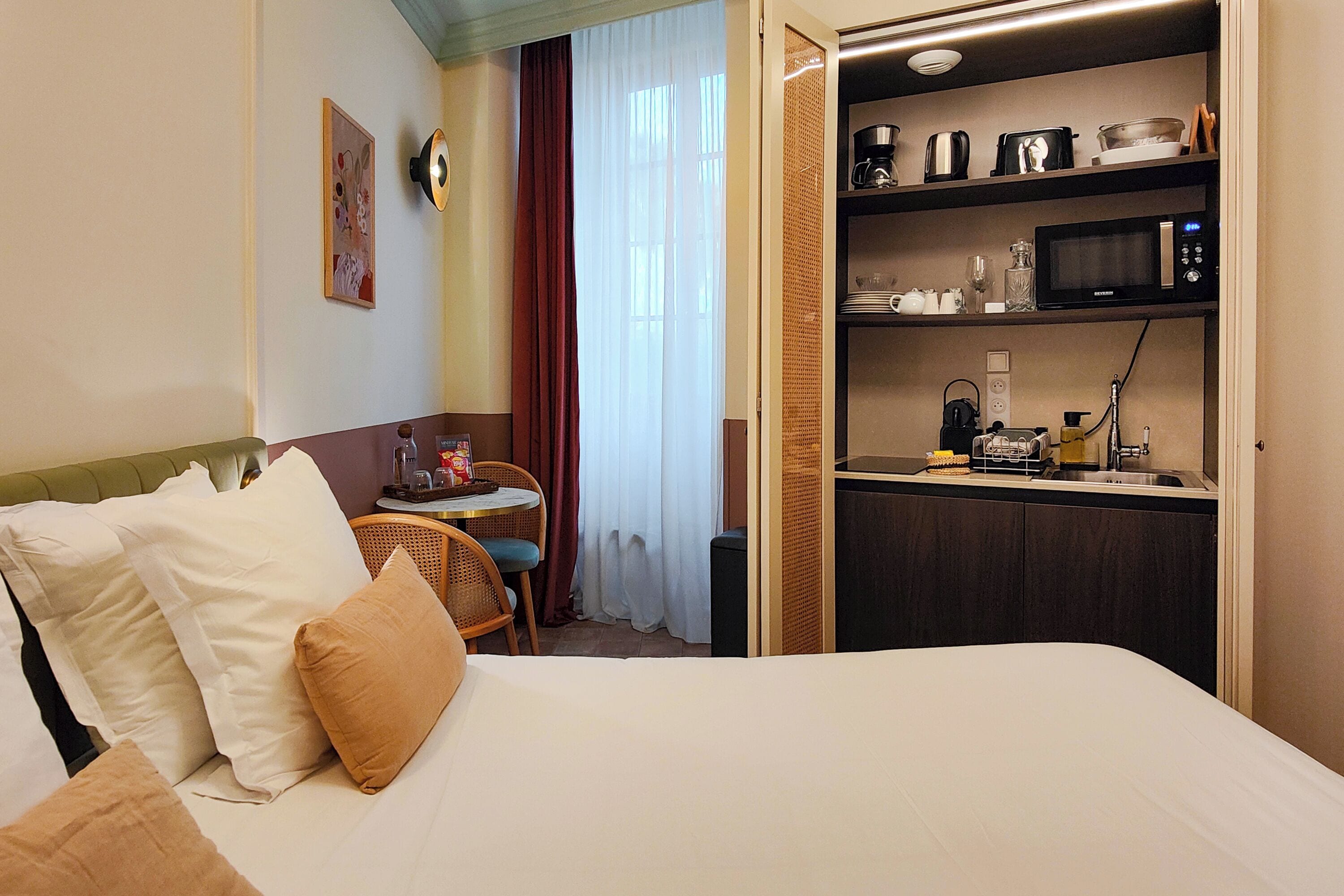 A hotel room with a bed, a small table with chairs, a kitchenette with a microwave, and a window with curtains.
