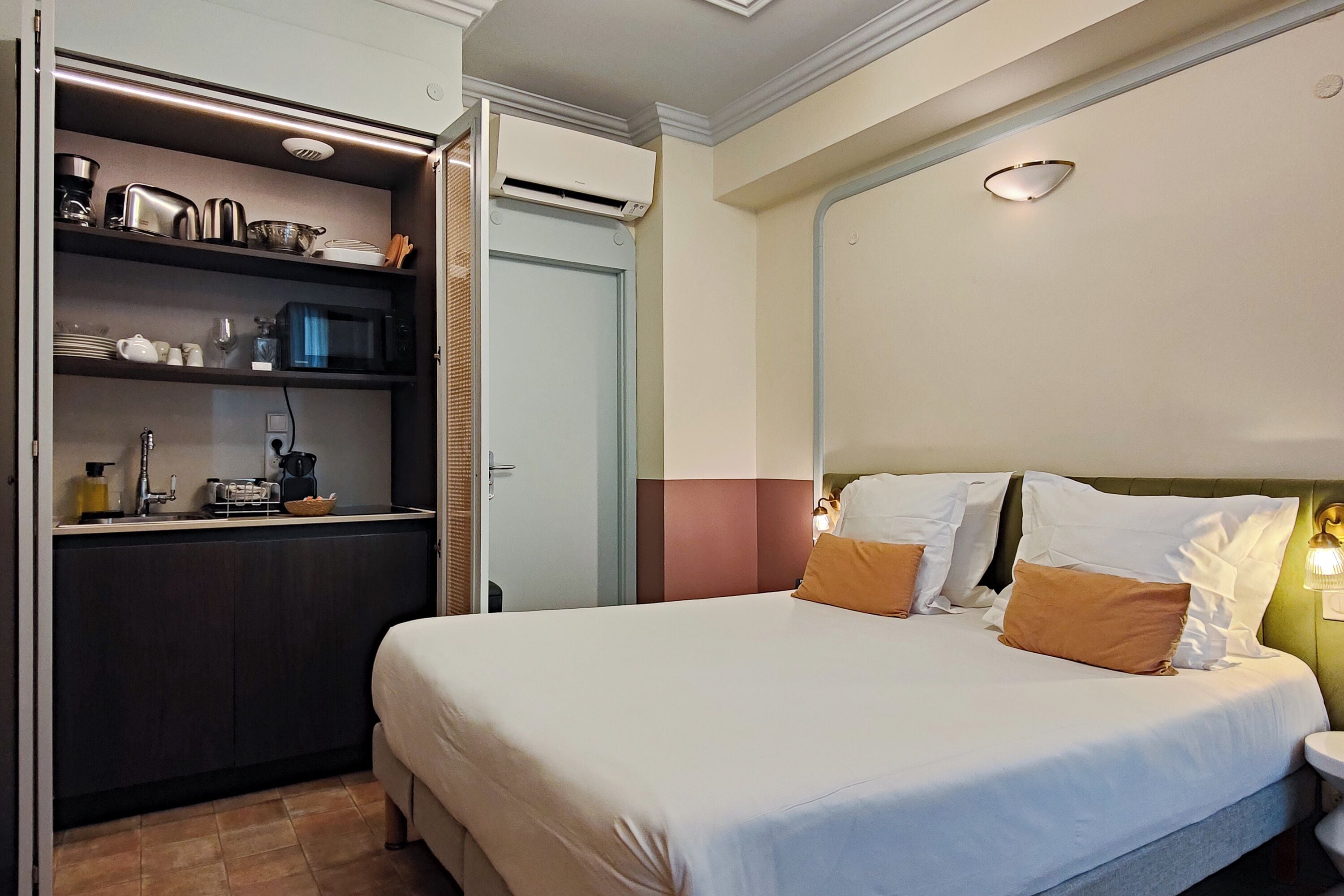 Una habitación de hotel con cama, mesita de noche y una kitchenette con microondas y otros electrodomésticos.