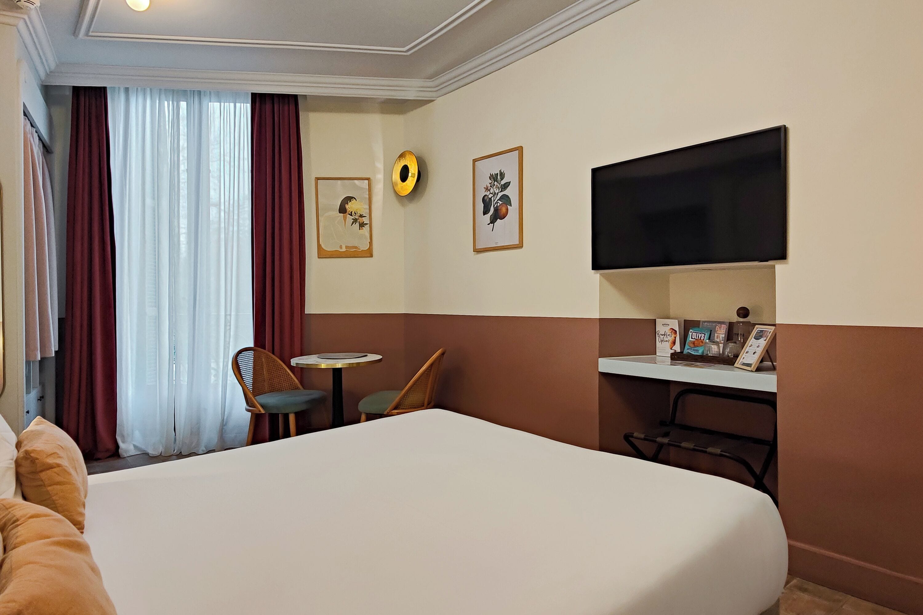 Hotellihuone, jossa on suuri sänky, seinälle asennettu televisio, pieni pöytä, kaksi tuolia ja ikkuna, jossa on verhot.