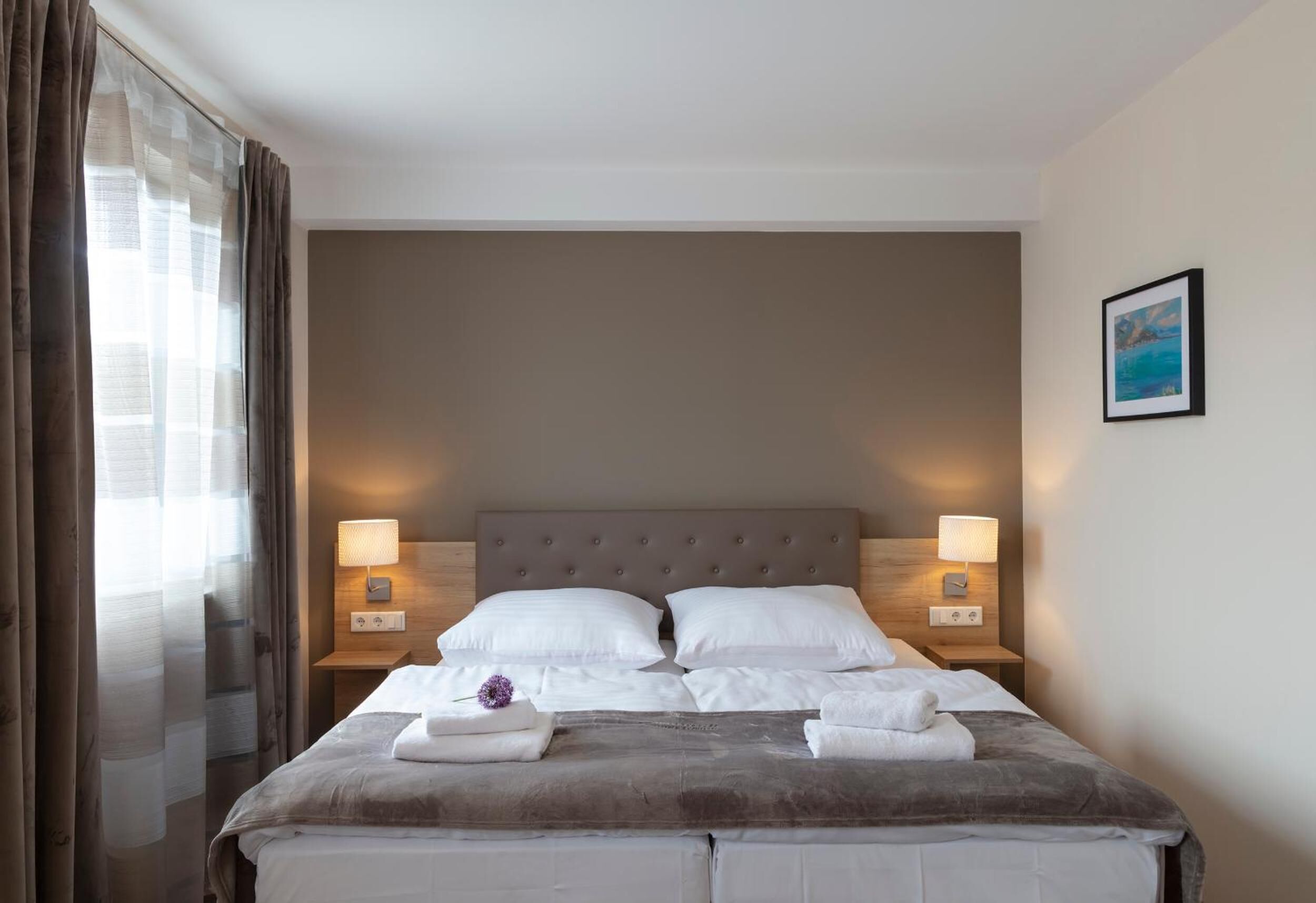 A neatly made bed with white linens and a gray headboard, two bedside lamps, and a framed picture on the wall.