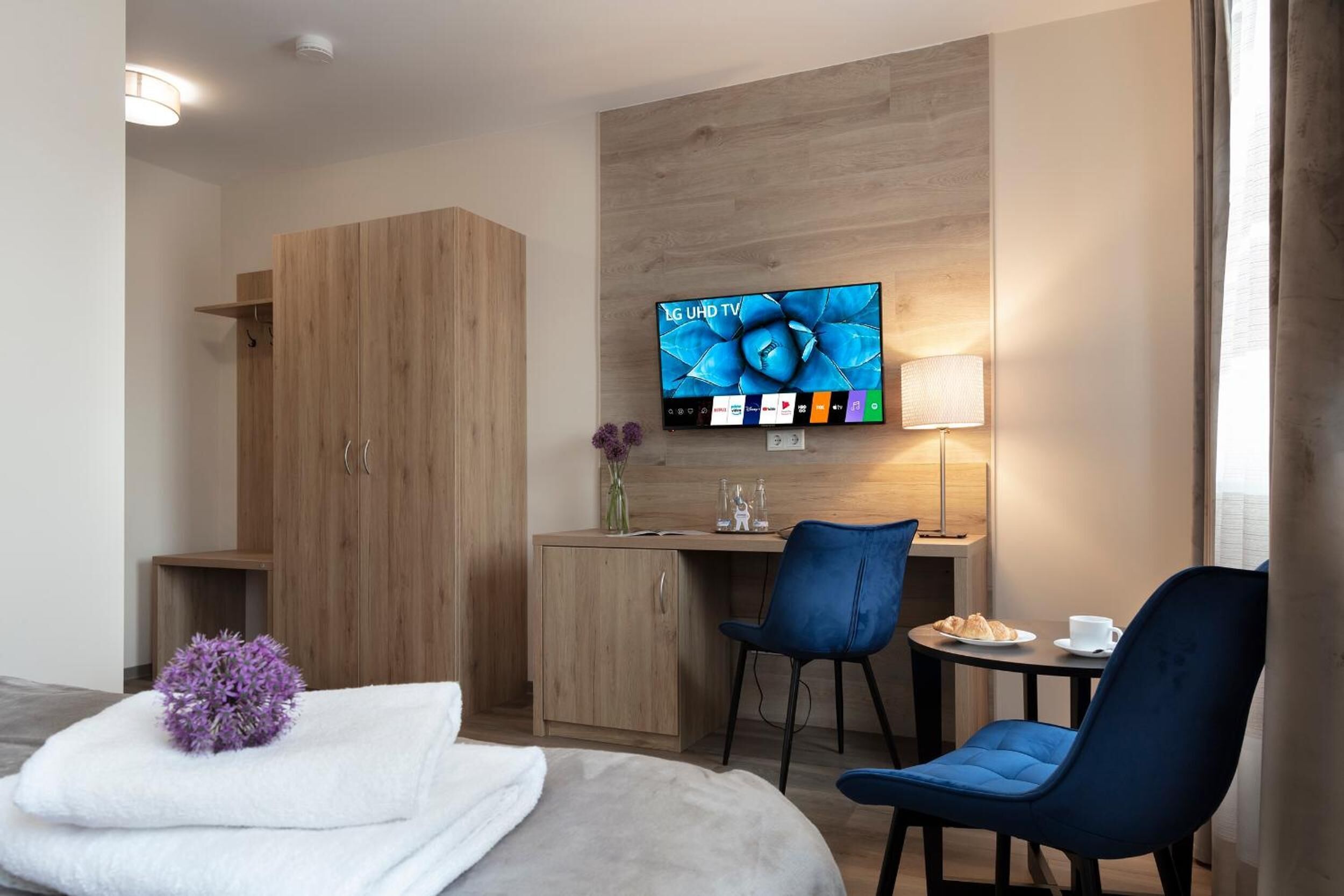 A hotel room with a bed, a desk with a chair, a TV on the wall, and a lamp.