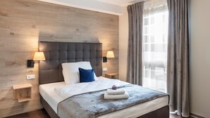 A hotel room with a wooden headboard, two bedside lamps, a bed with a white comforter and a blue pillow, a small wooden nightstand, and a window with curtains. - Dominik Motel (Paderborn)