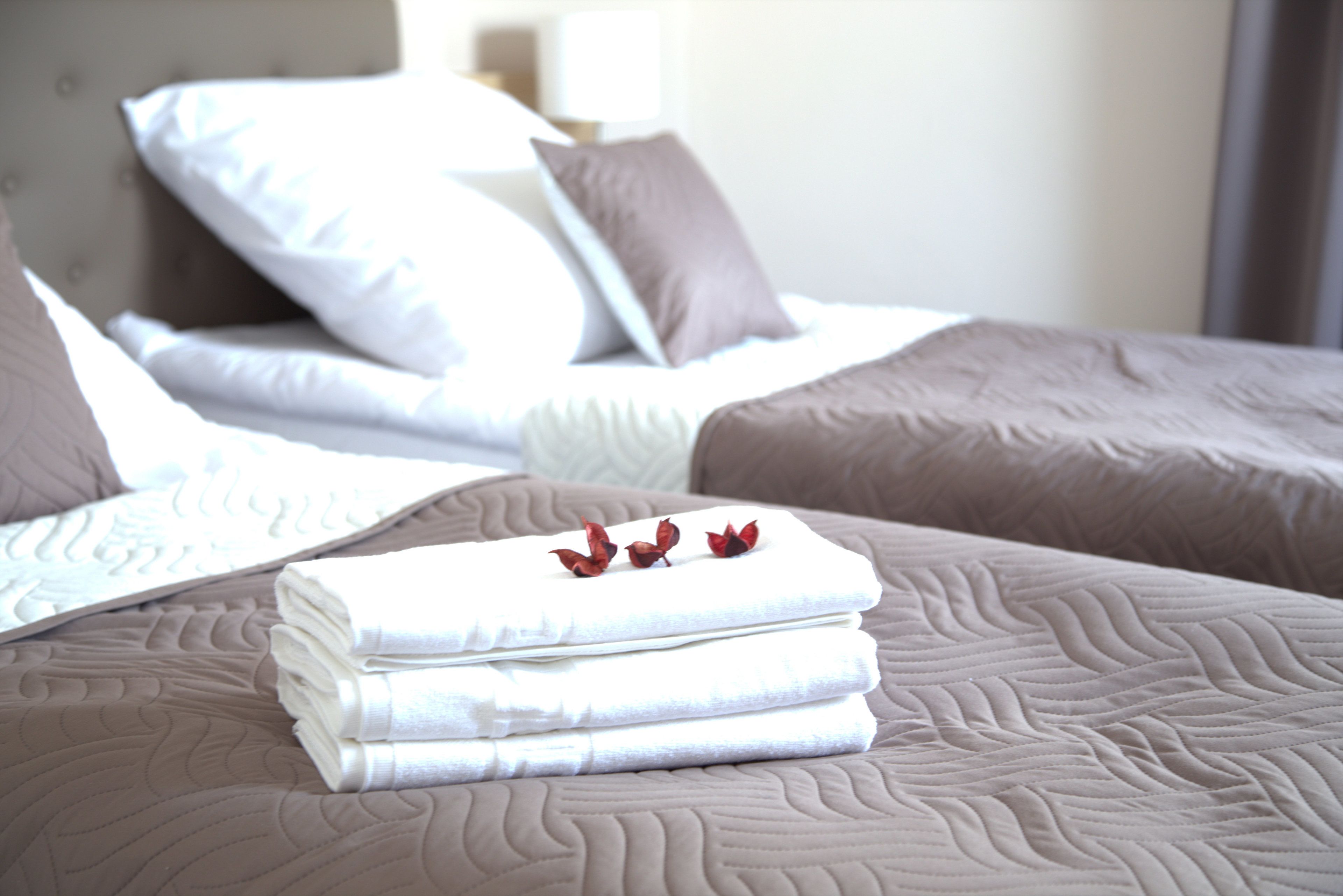 Una cama bien hecha con ropa de cama blanca y marrón, una pila de toallas dobladas y flores rojas decorativas sobre la cama.