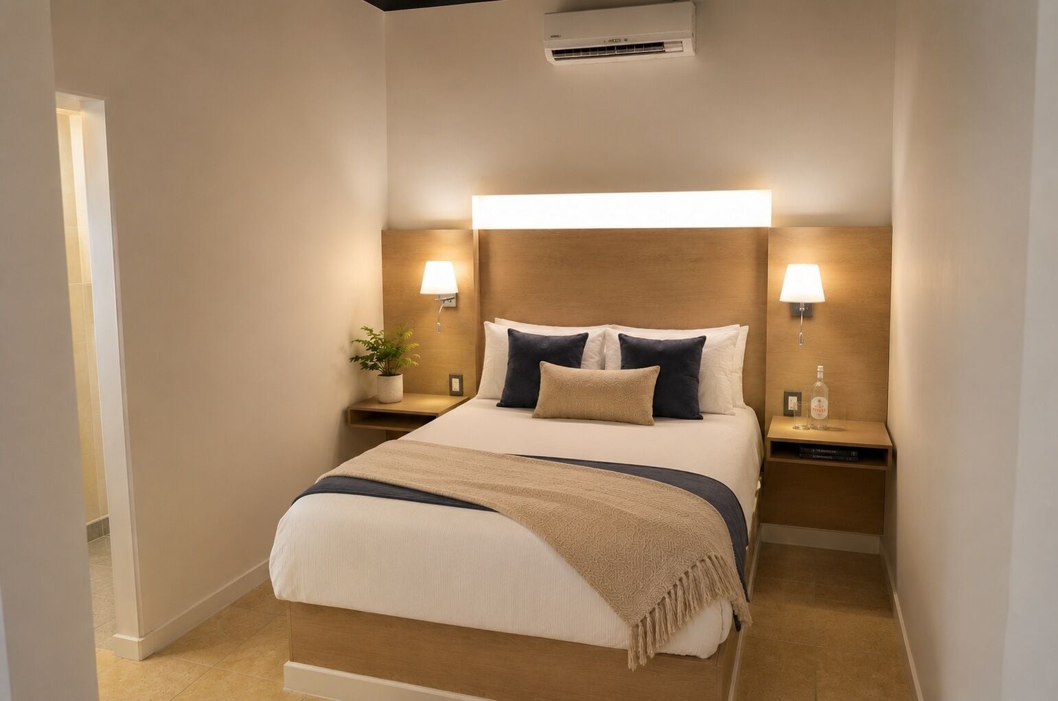 A hotel room with a large bed, bedside tables with lamps, a potted plant, and an air conditioning unit.