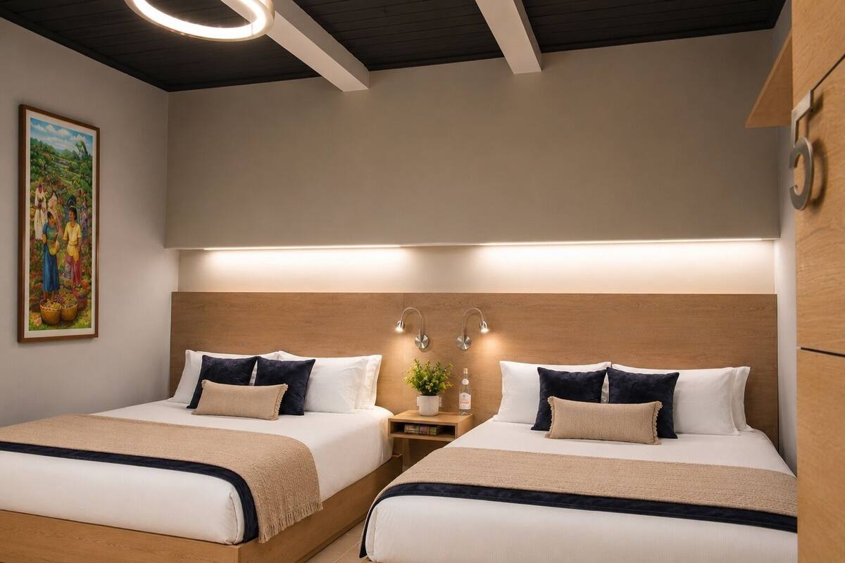 A hotel room with two beds, a wooden headboard, bedside lamps, a nightstand with a plant and a bottle, and a painting on the wall.
