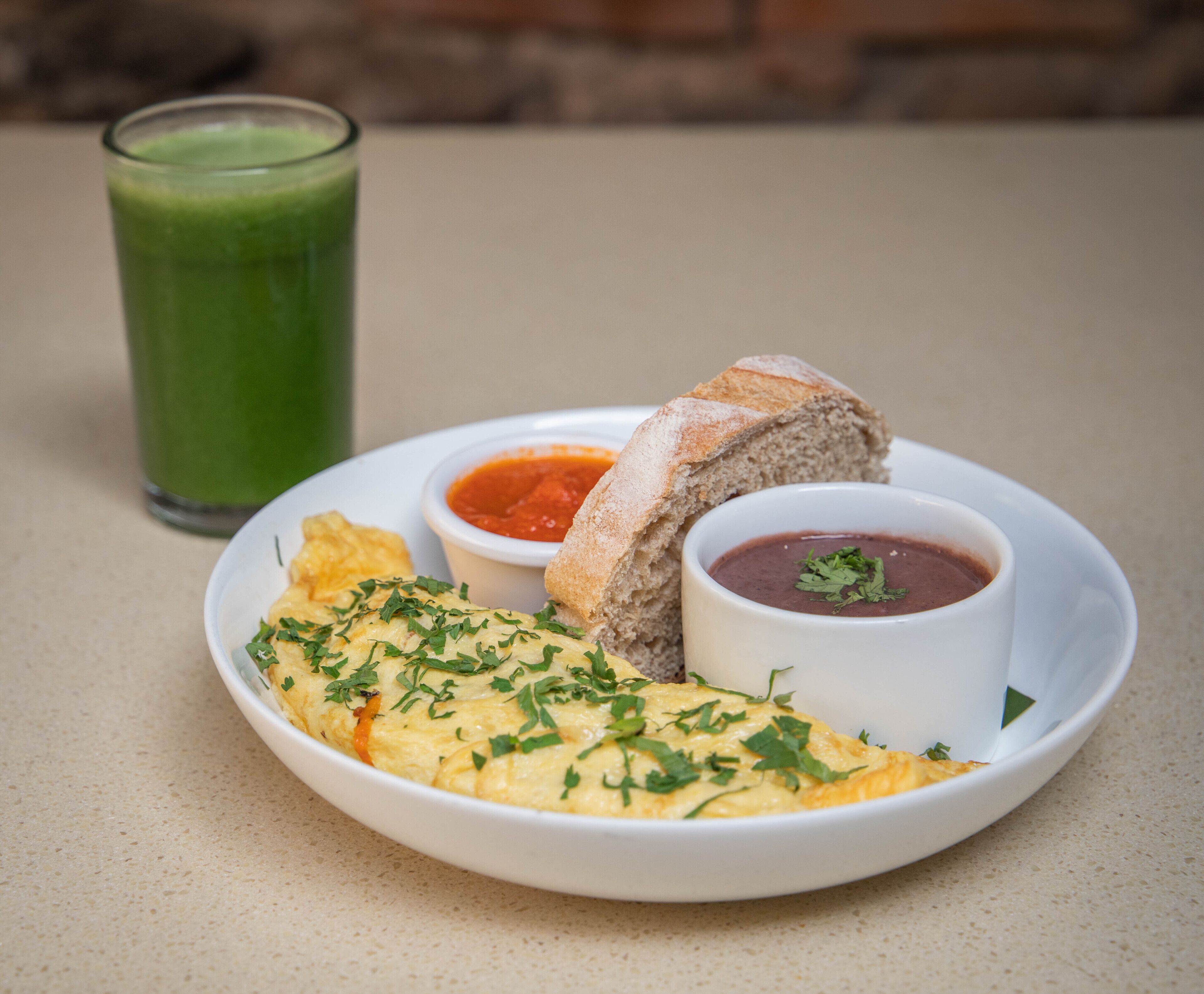 A plate with an omelette, bread, and two small bowls of sauce, accompanied by a glass of green juice.