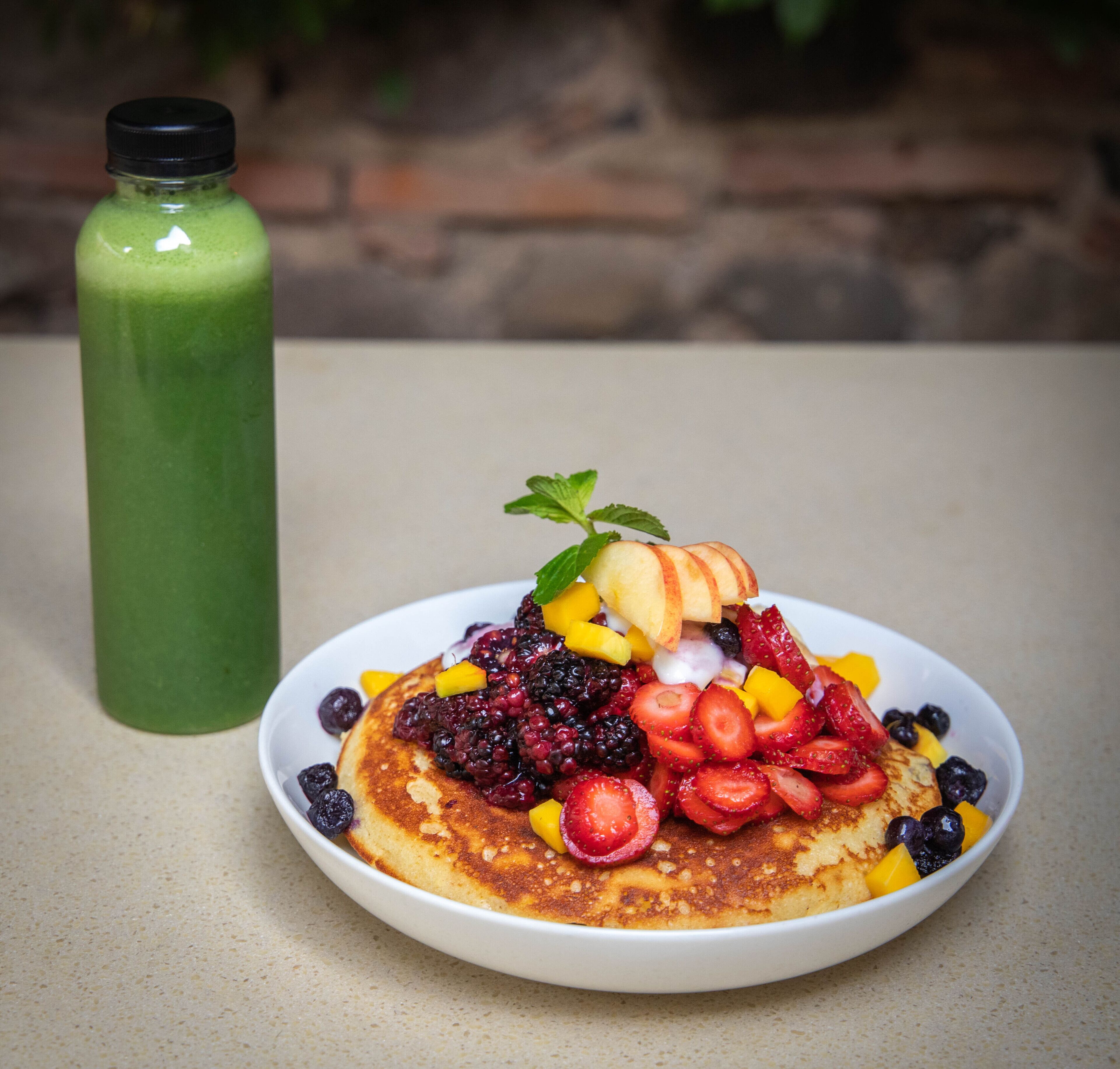 A plate of pancakes topped with various fruits and a green smoothie bottle beside it.