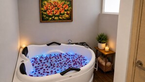 Een spa-ruimte met een bad gevuld met water en rozenblaadjes, een klein tafeltje met een plant en kaarsen, en een schilderij aan de muur.