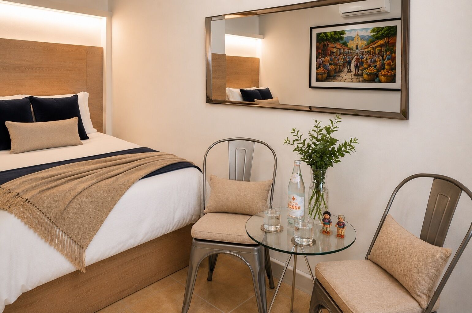 Una habitación de hotel con una cama, dos sillas, una mesita con un florero y una botella, y un cuadro en la pared.