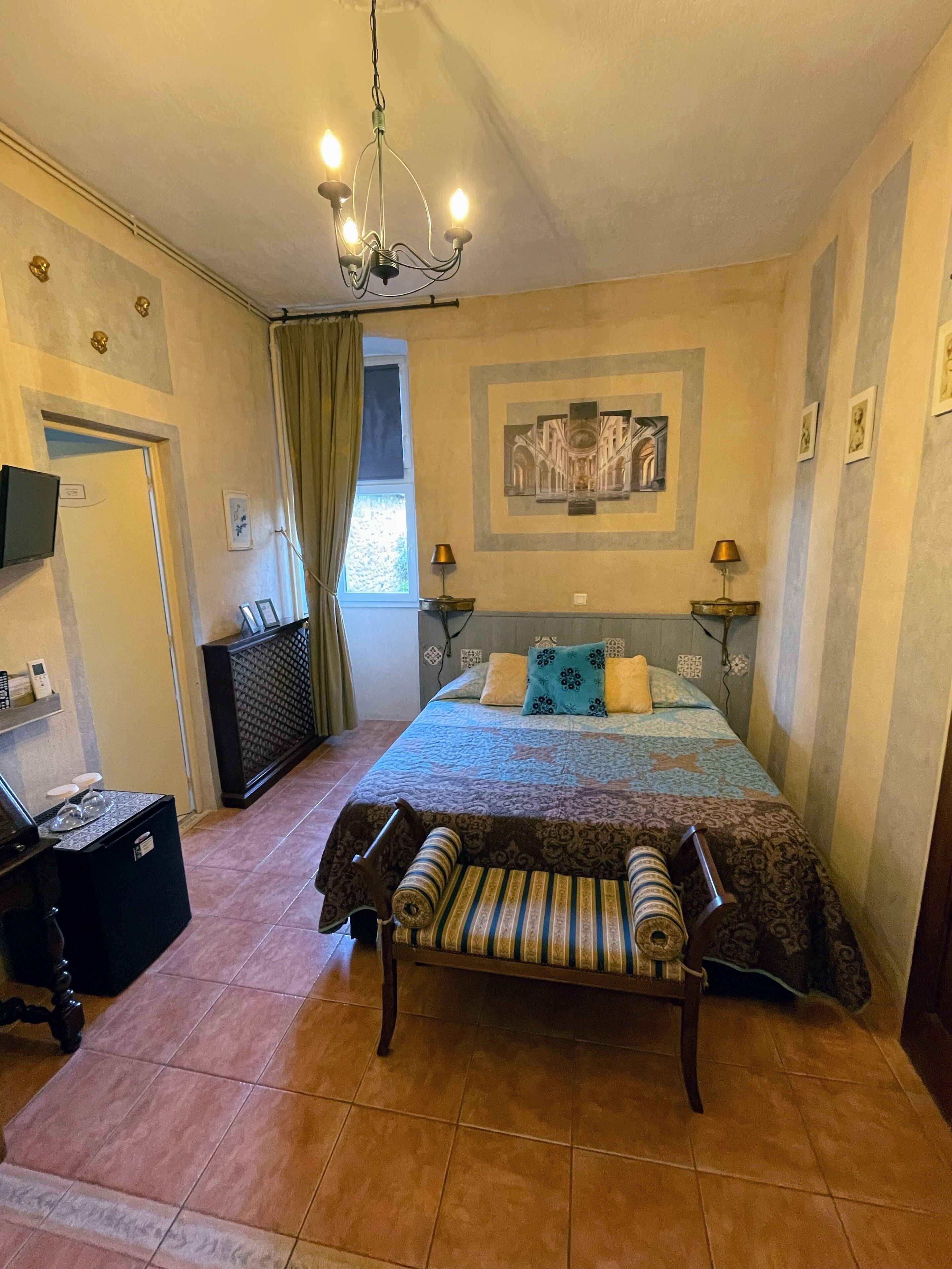 Ein Schlafzimmer mit einem Bett, einer Bank, einer Deckenleuchte, einem Gemälde und einem Fenster mit Vorhängen.