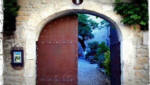 Un arco in pietra con una porta metallica decorativa, che dà accesso a un giardino con piante e un vialetto sterrato.