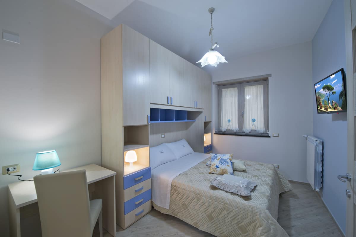 Quarto com cama, escrivaninha e cadeira. Há uma janela com cortinas, um radiador e uma televisão fixada na parede.