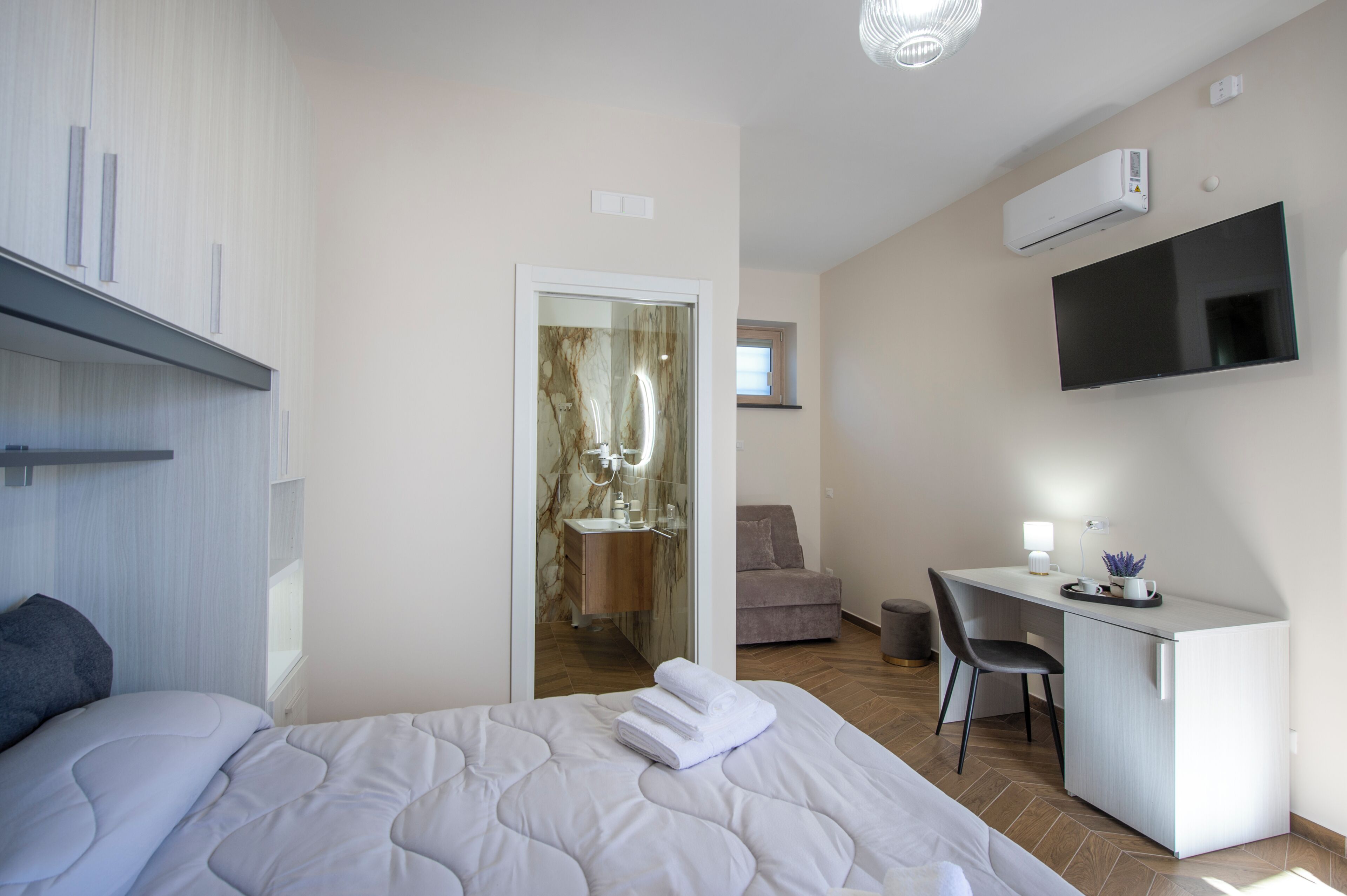 A modern hotel room with a bed, a desk, a chair, a TV, and an air conditioning unit.