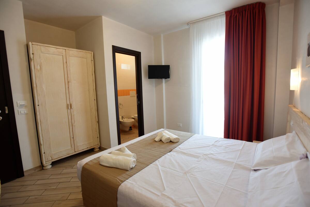 Una camera d'albergo con un letto, un armadio, una televisione e un bagno visibile attraverso una porta aperta.