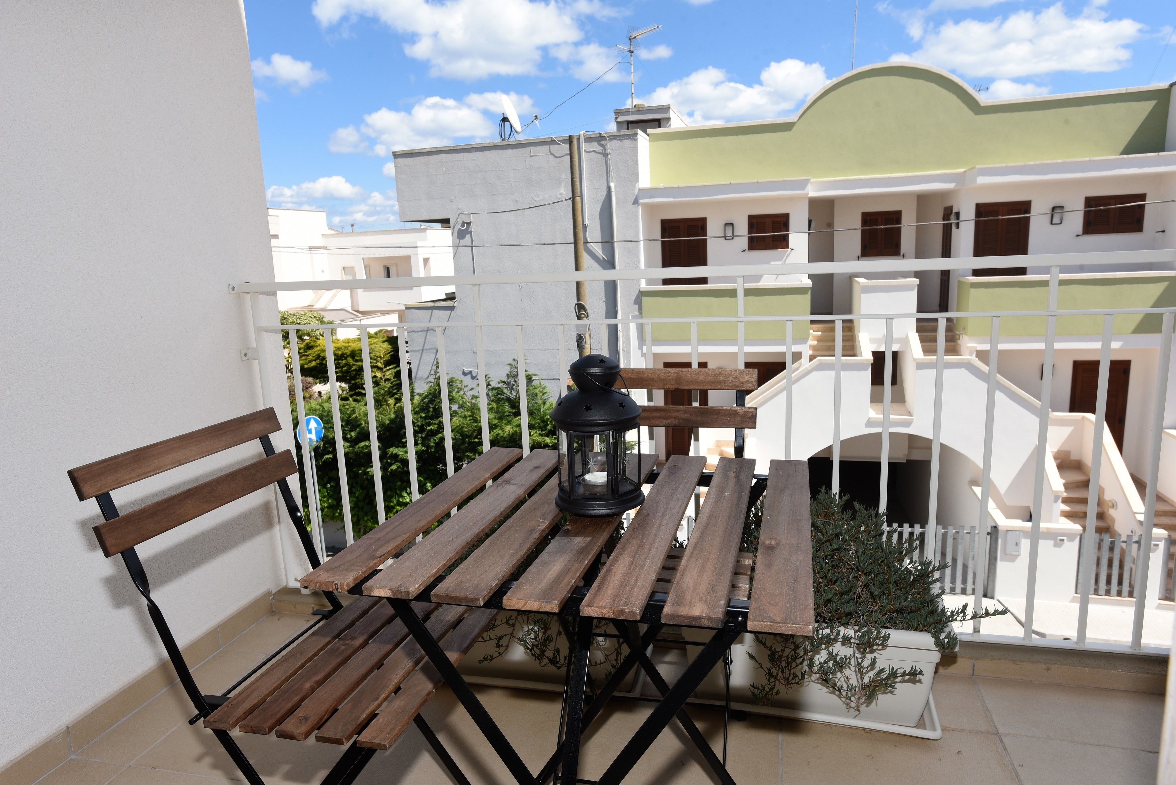 Un balcón con una mesa y bancos de madera, una planta en maceta y vistas a edificios vecinos.