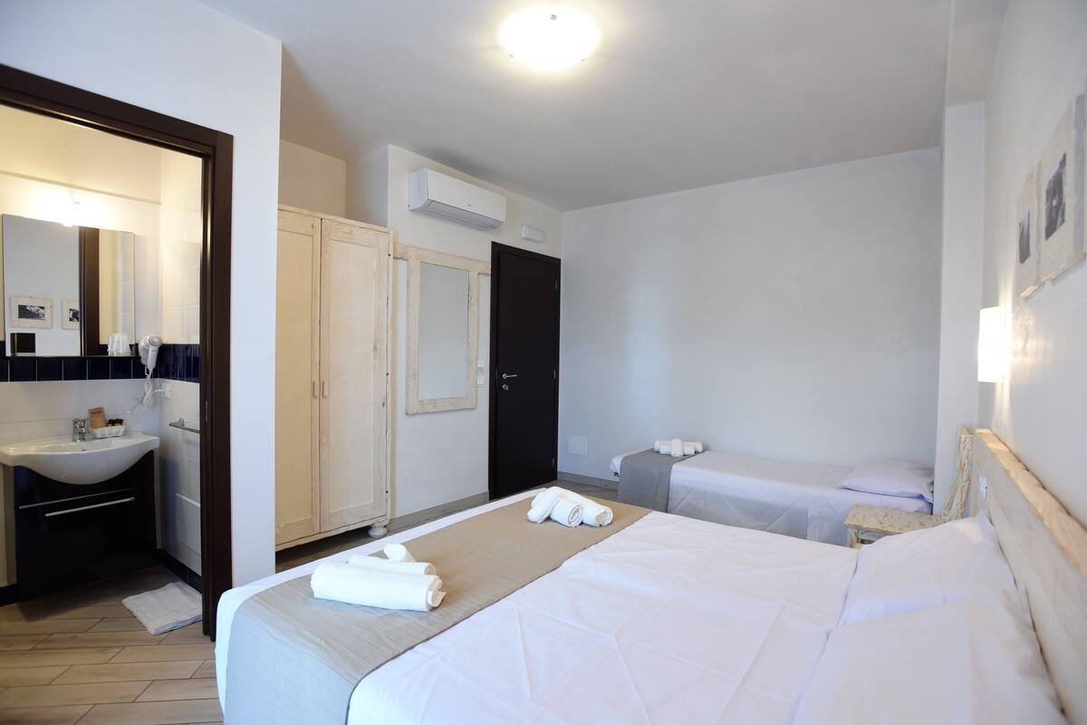 Camera d'albergo con un letto grande, un comodino, una piccola scrivania, una sedia, un armadio, uno specchio e un bagno con lavandino e porta asciugamani.