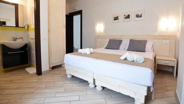 Une chambre à coucher avec un lit en bois, une table de chevet, une commode, un miroir et une salle de bain équipée d’un lavabo et d’un porte-serviettes.