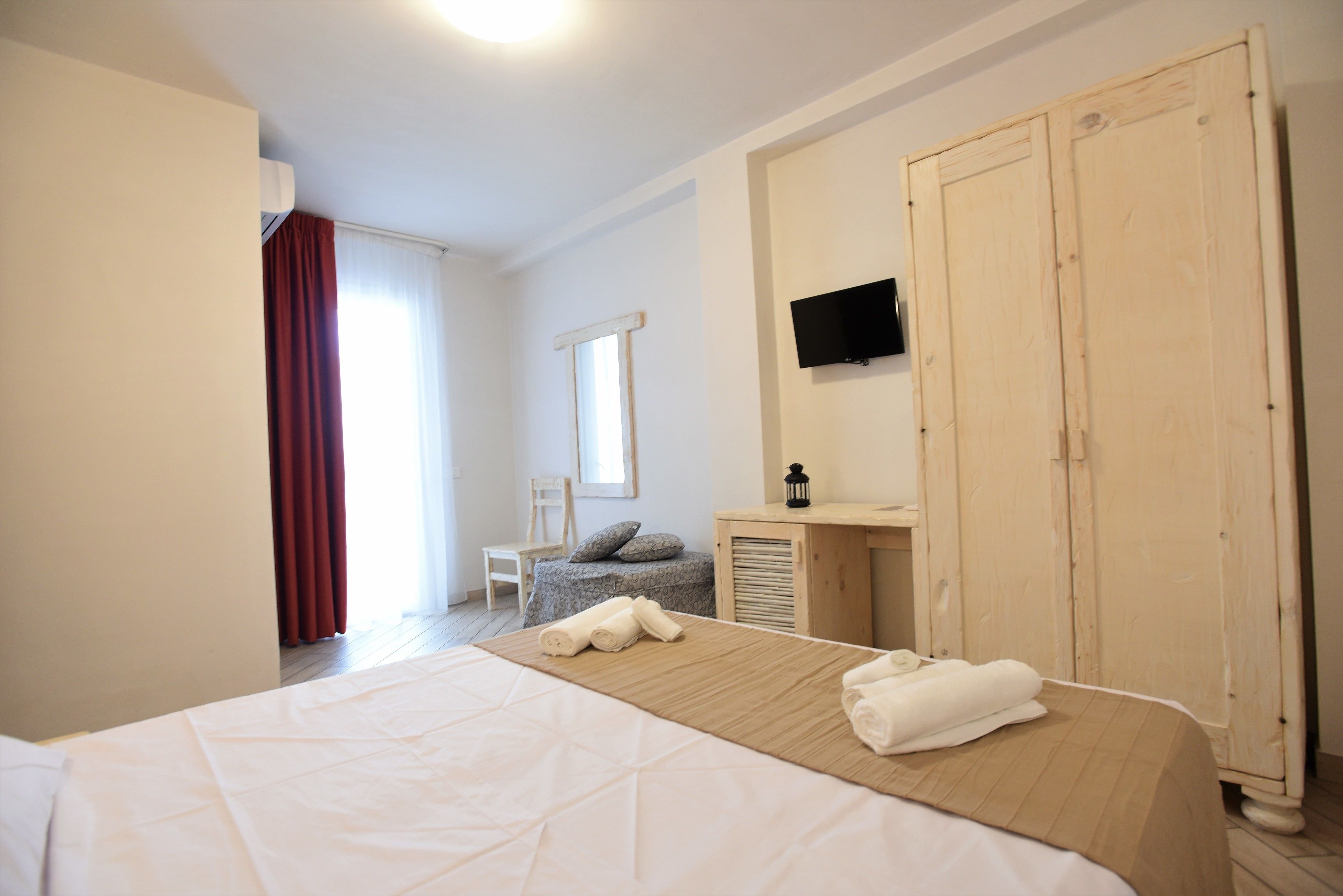 A hotel room with a large bed, a wooden wardrobe, a television mounted on the wall, and a window with curtains.