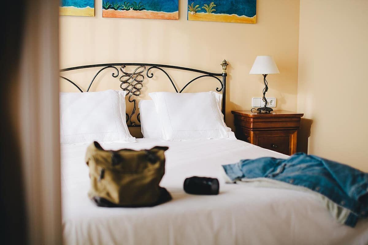 Un lit avec du linge de lit blanc, une tête de lit en métal, une table de chevet avec une lampe, et un chat sur le lit.